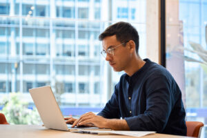 A man wearing glasses and a dark shirt works on a laptop at a desk in a modern office with large windows and an urban cityscape in the background.