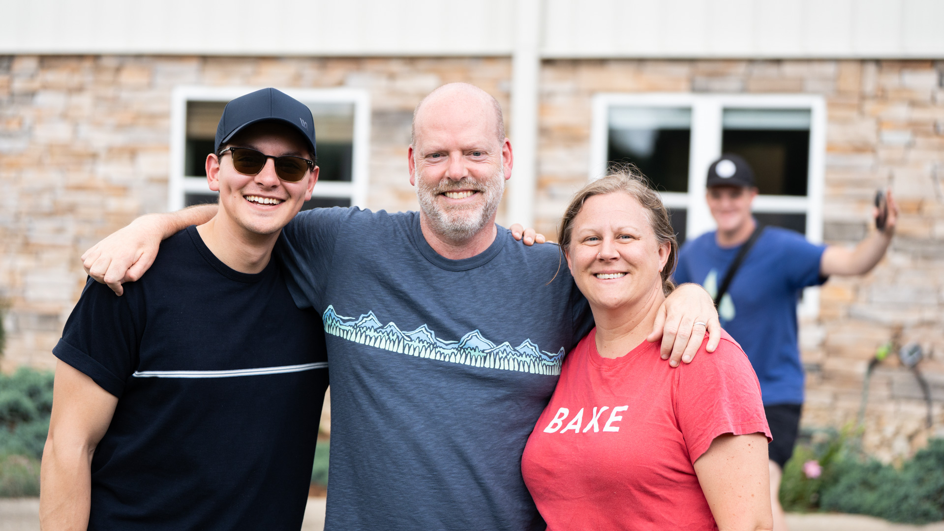 Three people stand arm-in-arm smiling at the camera outdoors, with a stone building and windows behind them. A fourth person in the background waves and smiles—a joyful moment captured through our photography & videography services.