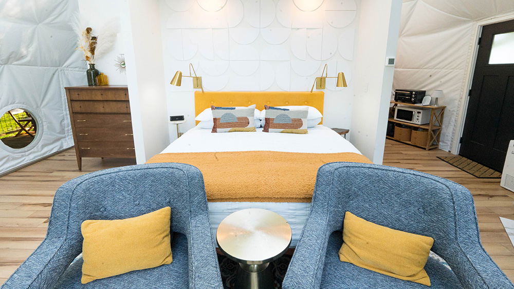 Modern bedroom with a yellow-accented bed, blue armchairs, and a kitchenette—perfect for photography & videography services in a bright, airy space with wood floors and white walls.
