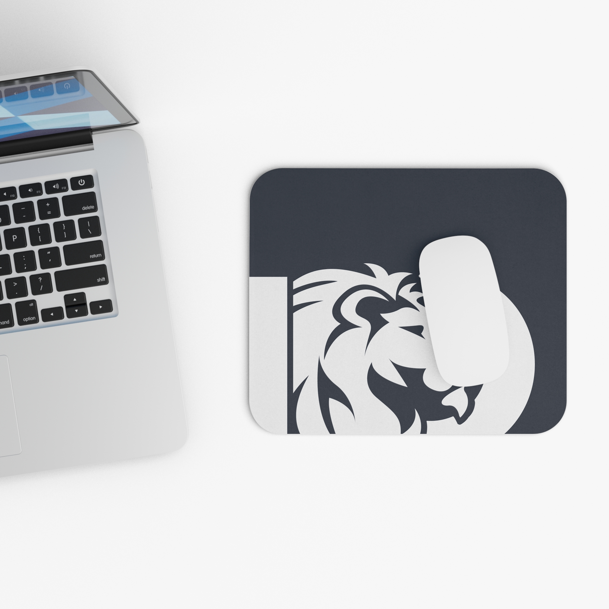 A silver laptop sits beside a Lion Logo Mouse Pad — Minimal Dark Desk Mat for Office & Gaming, with a white wireless mouse on top, all on a white surface.