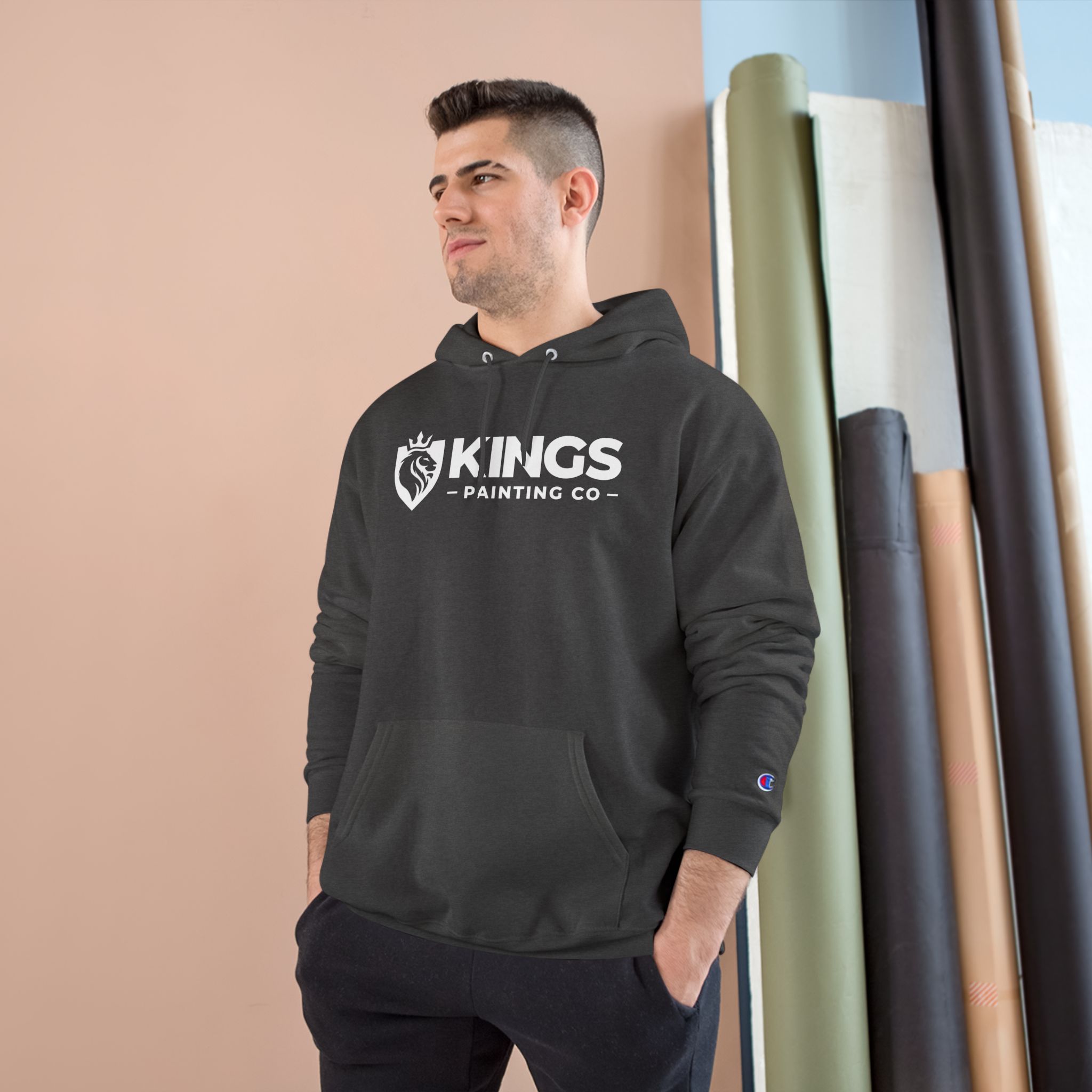 A man stands in front of a beige wall and rolls of fabric, looking to the side with his hands in his pockets, wearing the Kings Painting Co. Logo Hoodie by Champion®.