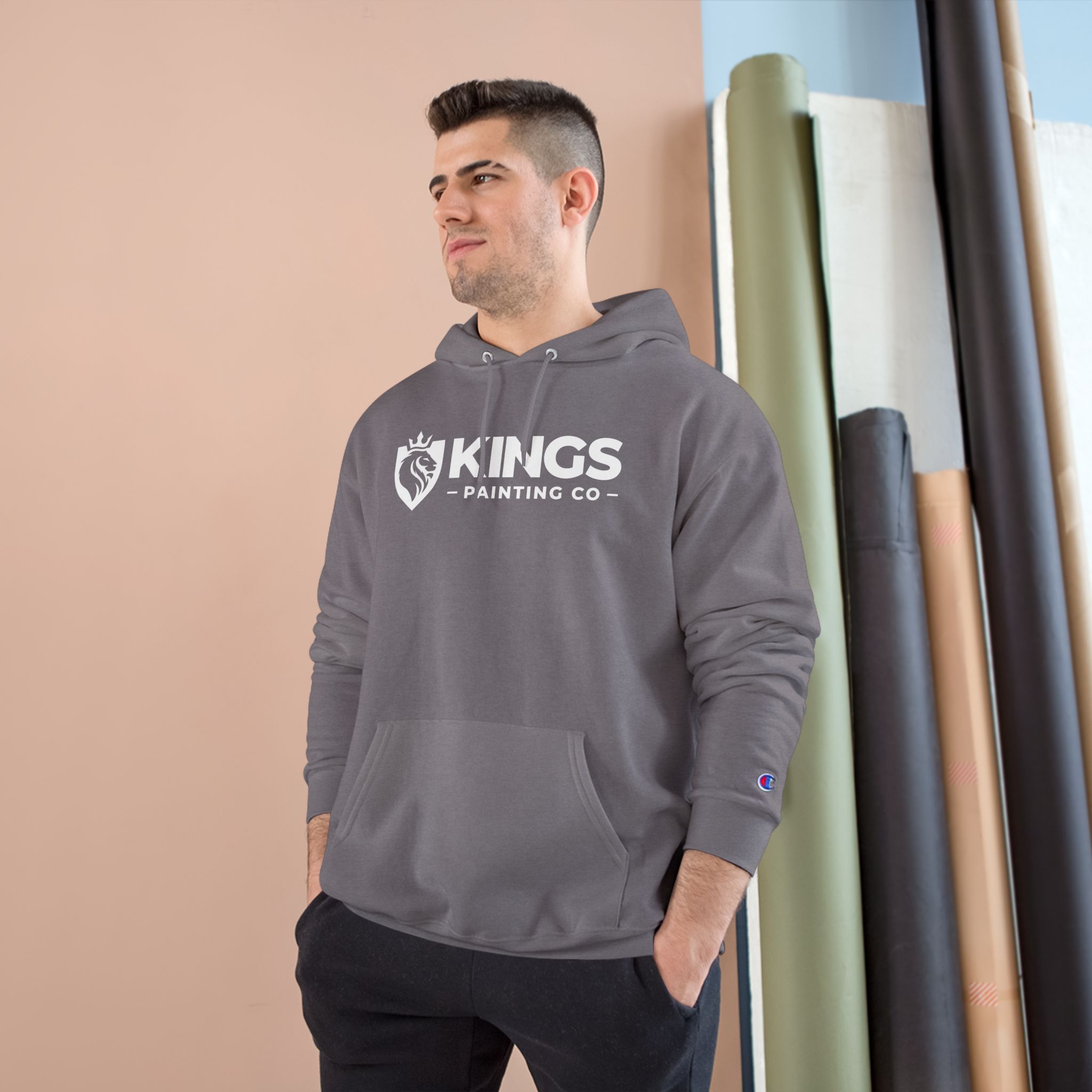 A man wearing the Kings Painting Co. Logo Hoodie by Champion® stands in front of a tan and blue background, with rolled-up papers or fabrics leaning against the wall.