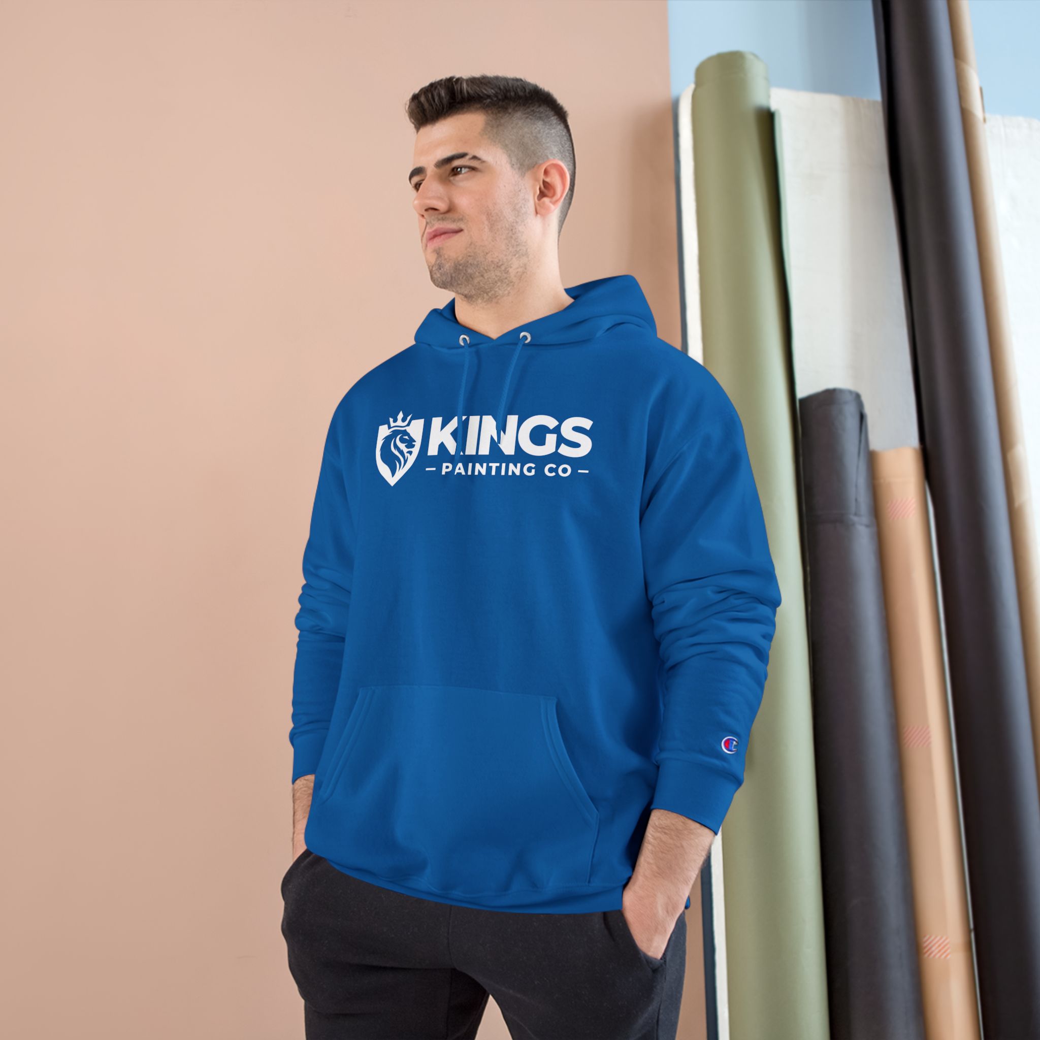 A man stands with his hands in his pockets, looking to the side, while wearing the Kings Painting Co. Logo Hoodie | Champion®. In the background, rolls of paper or fabric are stacked vertically against a wall.