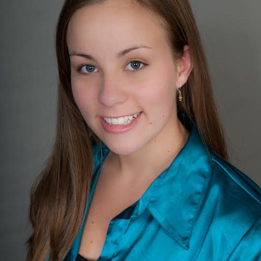 A woman with long brown hair smiles warmly at the camera, exuding the creativity of web development and design. She is wearing a turquoise satin blouse and gold earrings. The neutral gray background enhances this professional portrait setting, reminiscent of Ashevilles vibrant artistry.